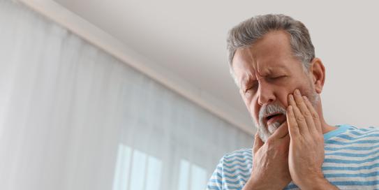 Älterer Mann mit Bart hält sich schmerzverzerrt die Wange, möglicherweise wegen Zahnschmerzen.