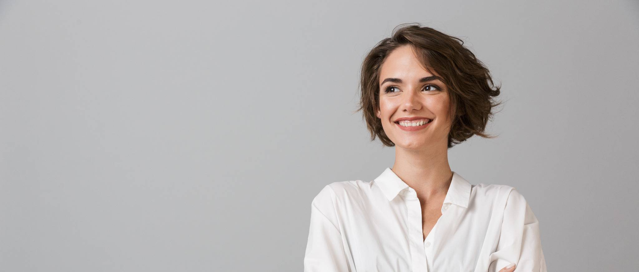 Junge Frau mit kurzem braunen Haar lächelt und verschränkt die Arme, vor grauem Hintergrund.