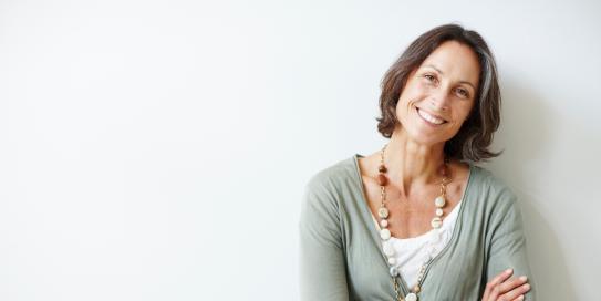 Reifere Frau mit halblangen braunen Haaren steht lächelnd mit verschränkten Armen vor einer weißen Wand, trägt eine lange Kette und einen hellgrünen Cardigan.