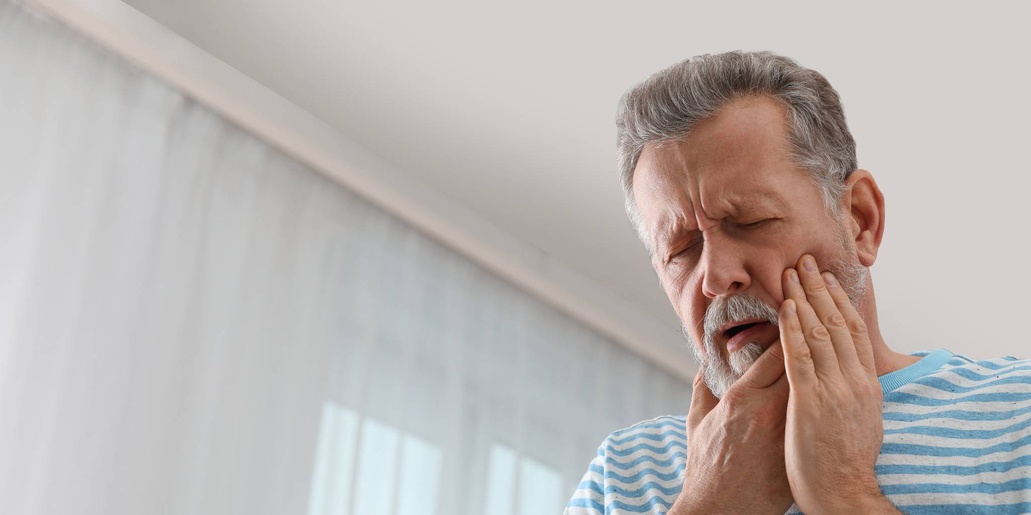Älterer Mann mit Bart hält sich schmerzverzerrt die Wange, möglicherweise wegen Zahnschmerzen.