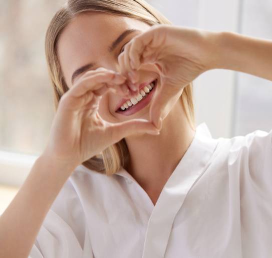 Junge Frau in weißem Hemd formt mit den Händen ein Herz vor ihrem Gesicht und lächelt.